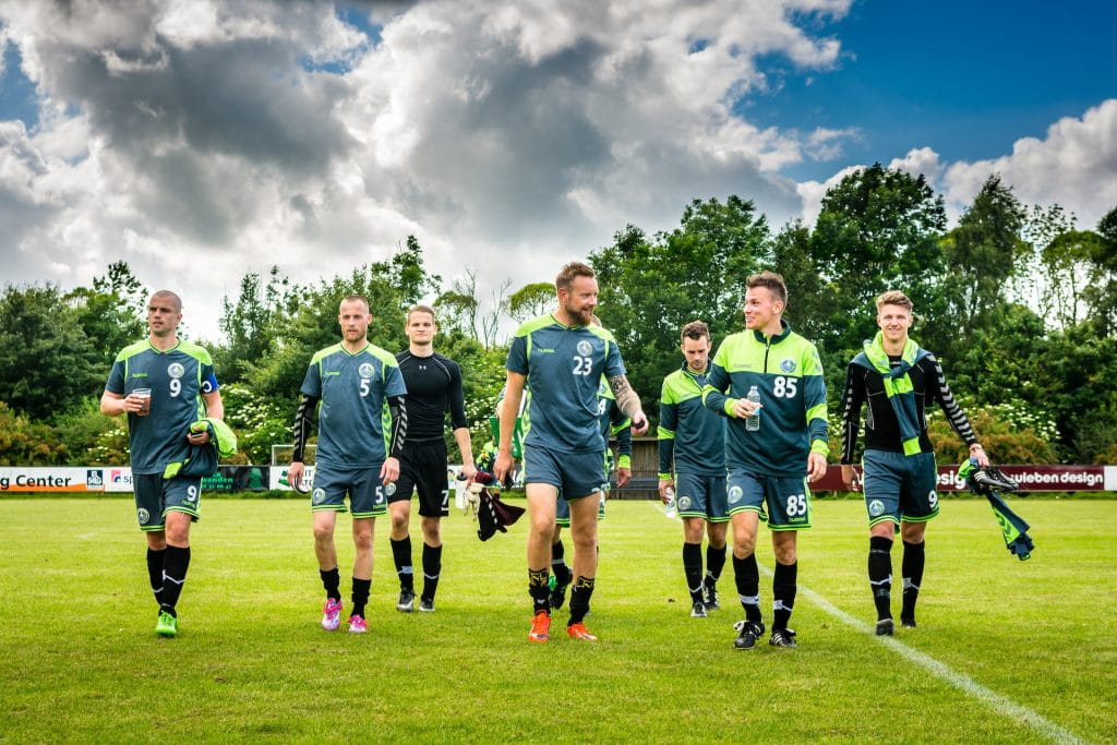 Seven male soccer players walking towards the camera on an outdoor pitch after finishing a game.