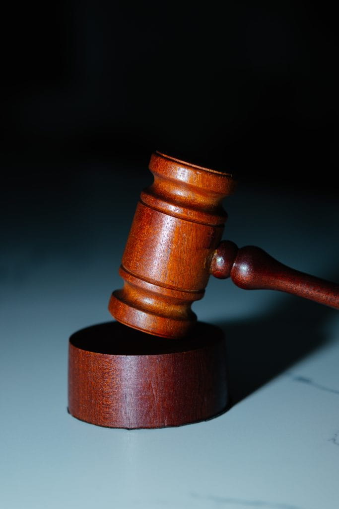 A brown-stained wooden gavel rests on a greyish-blue surface. The shadow in the background gives an ominous and serious feel.