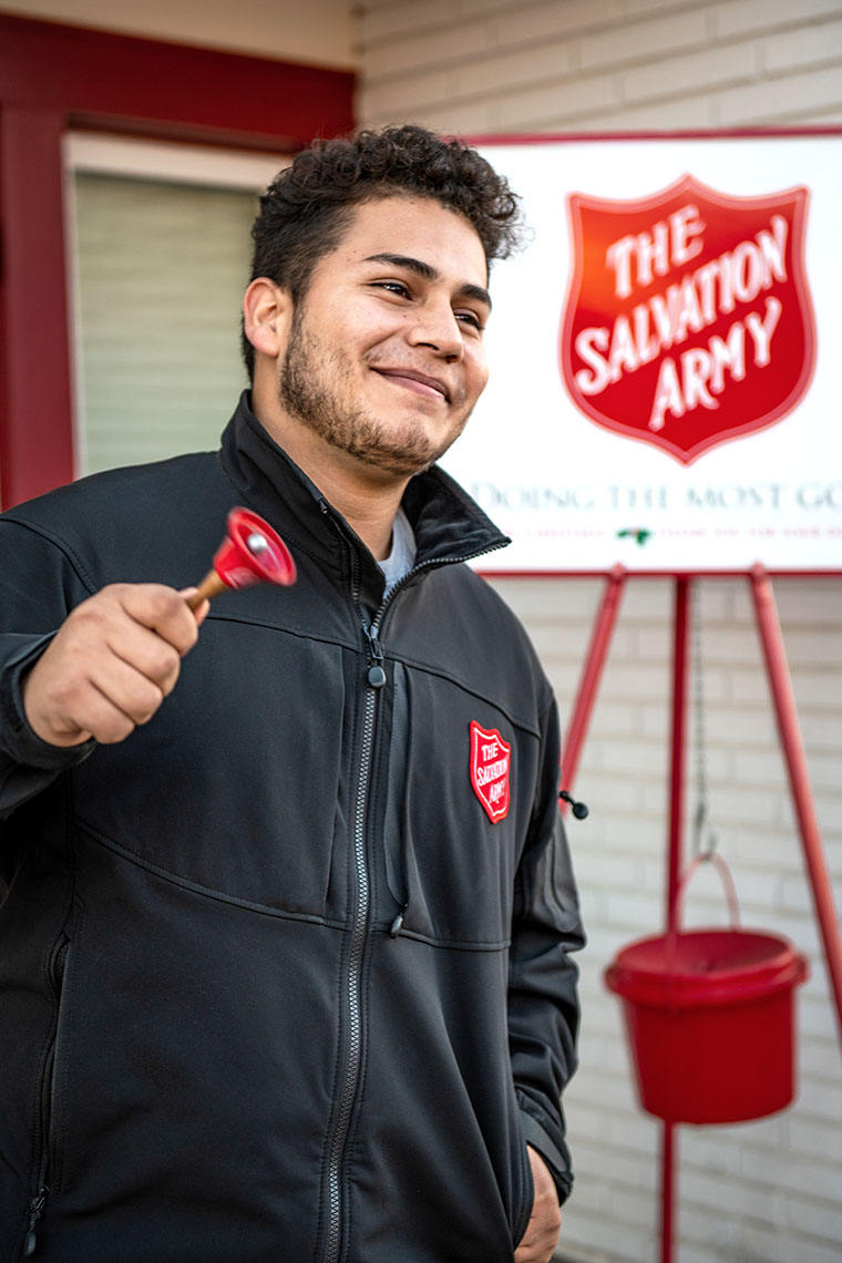 A young man rings a small red bell. He wears a black jacket with a Salvation Army badge sewn on its front. A Salvation Army sign with a collection bucket hanging from it is behind him.