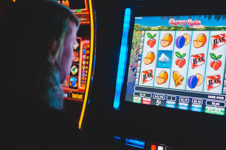 A person with long hair stares at the flat screen of an electronic gambling machine. Fruit, bells and other colourful icons are in columns on the screen. 