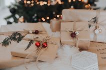 Brown paper-wrapped gifts, tied with jute string and garlanded with bells and pine sprigs are piled on top of each other. A Christmas tree with lights and decorations is blurred in the background.
