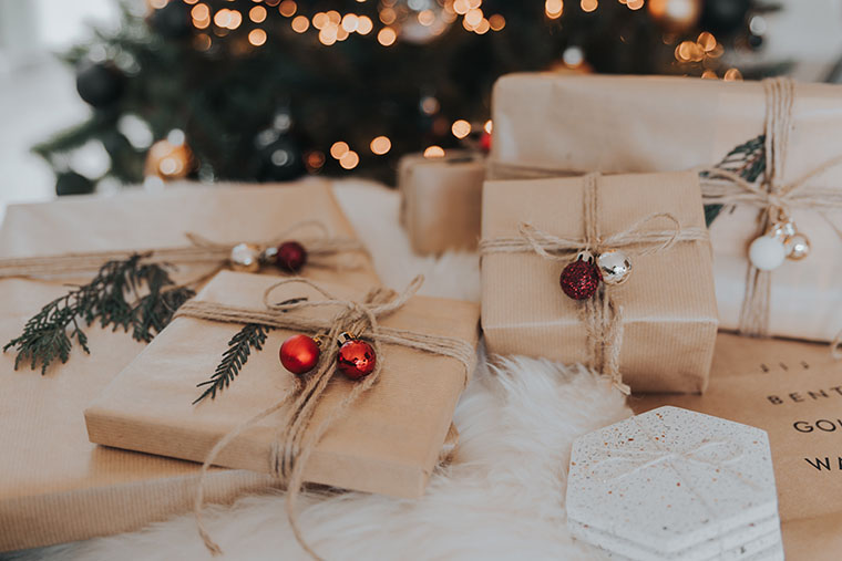 Brown paper-wrapped gifts, tied with jute string and garlanded with bells and pine sprigs are piled on top of each other. A Christmas tree with lights and decorations is blurred in the background. 