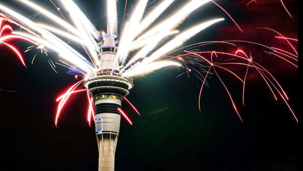 Sky Tower, by the casino in Auckland, has fireworks spurting out of its top in all directions. The sky is dark and some of the fireworks look neon-coloured.