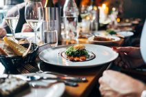 A long wooden table is full to the brim with plates, glasses and cutlery. A pale blue ceramic dinner plate hosts an expensive-looking modern course of fish and salad. A manicured hand is touching the plate and a basket of bread is behind it.