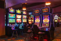 Gaming machines with colourful screens sit with empty chairs in front of them in a gambling lounge. The room is dimly lit, making the screens seem even brighter.