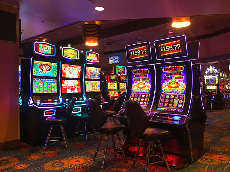 Gaming machines with colourful screens sit with empty chairs in front of them in a gambling lounge. The room is dimly lit, making the screens seem even brighter.