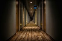 A long hallway with chicly rough wooden floorboards continues on into the distance. On either side grey walls with brown hotel doors lead off it.