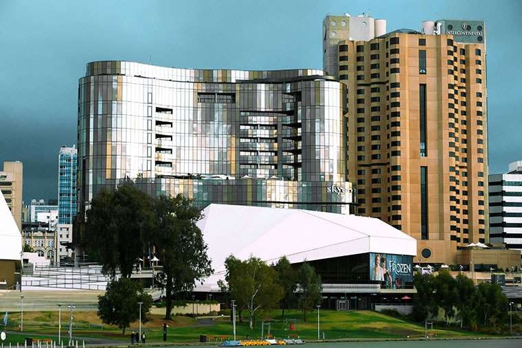 SkyCity Adelaide’s shiny silver and curved building is sitting next a large taller brown building. A blue and grey sky is clear behind them. 